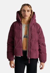 Burgundowa kurtka puchowa z kapturem, z zapinaniem na zamek, elastycznymi mankietami i pikowanym wzorem. Noszona na szarym T-shircie i czarnych legginsach.