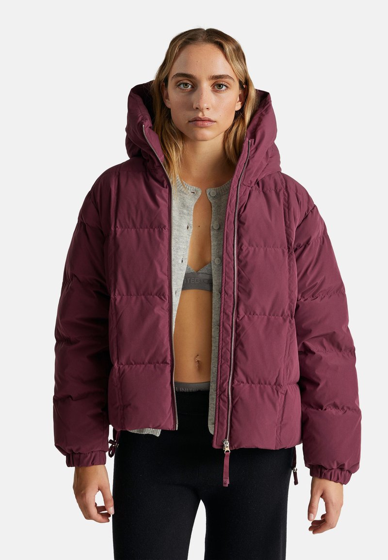 Burgundowa kurtka puchowa z kapturem, z zapinaniem na zamek, elastycznymi mankietami i pikowanym wzorem. Noszona na szarym T-shircie i czarnych legginsach.