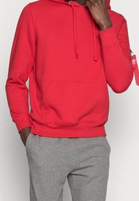 Felpa rossa con cappuccio, tasca a marsupio, polsini e orlo a coste. Presenta una tasca con zip sulla manica sinistra. Abbinata a pantaloni della tuta grigi.