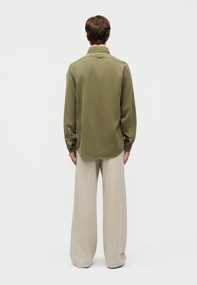 Άνδρας που στέκεται γυρισμένος με την πλάτη, φορώντας πουκάμισο με μακριά μανίκια σε πράσινο ελιάς και φαρδιά μπεζ παντελόνια, μπροστά σε απλό φόντο.