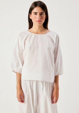 Donna con capelli lunghi e scuri che indossa una maglia bianca larga con maniche a tre quarti e pantaloni bianchi abbinati su uno sfondo semplice.
