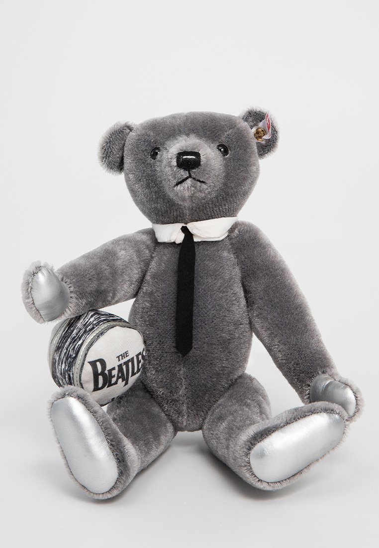 Grauem Plüschbär mit einem weißen Hemd und schwarzer Krawatte. Silberne Akzente an den Pfoten, die einen runden Gegenstand halten, der mit "The Beatles" beschriftet ist.