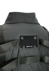 Chaqueta acolchada gris con cuello alto, correas horizontales y una etiqueta negra con ojales. Textura suave y ligeramente brillante.