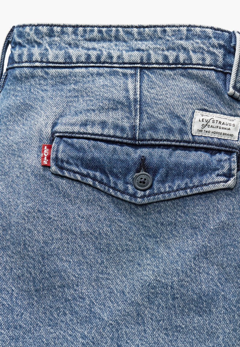 Blaue Denim-Jeans mit einer Gesäßtasche, die mit einem einzelnen schwarzen Knopf und einem Levis-Label mit weißem Text versehen ist. Der Stoff hat eine ausgewaschene Textur.