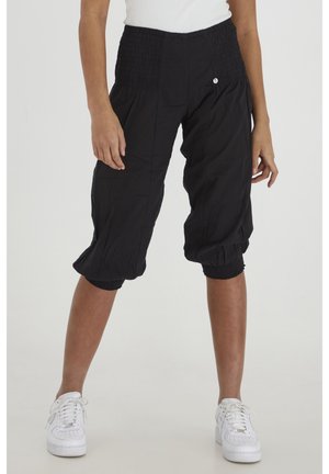 Schwarze, verkürzte Hosen mit elastischem Bund und Bündchen, getragen mit weißen Sneakers und einem weißen Oberteil an einer stehenden Person.