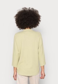 Pull vert clair texturé avec manches 3/4, encolure ronde et ourlet arrondi. Porté avec un pantalon clair, affichant une coupe décontractée.