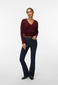 Femme debout, une main dans la poche avant, portant un pull col en V bordeaux, un jean évasé bleu foncé et des chaussures plates noires.