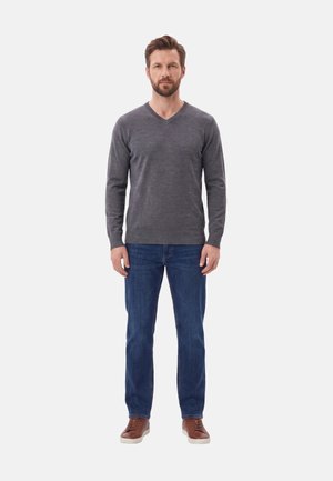 Uomo in piedi indossa un maglione grigio a V, jeans blu e scarpe da ginnastica marroni su uno sfondo chiaro e uniforme.