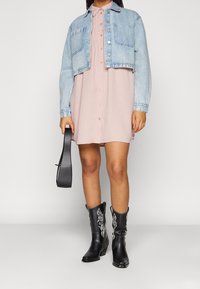 Jeansjacke in Hellblau, die über einem pinken Hemdkleid getragen wird, kombiniert mit schwarzen Cowboystiefeln mit weißer Stickerei und einer schwarzen Tasche.