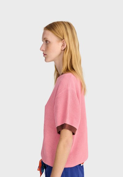 Femme aux longs cheveux blonds portant un pull en tricot rose à manches courtes avec des poignets marron et un pantalon bleu, tournée vers la gauche sur un fond uni.