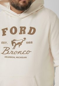 Beżowa bluza z kapturem z kieszenią typu kangur, z brązowymi napisami: "FORD" na górze oraz "Bronco Dearborn, Michigan" poniżej, a także grafiką galopującego konia.
