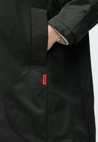 Mână în buzunarul unei jachete negre cu un model circular subtil și eticheta de țesătură roșie MAX&Co pe cusătura laterală.