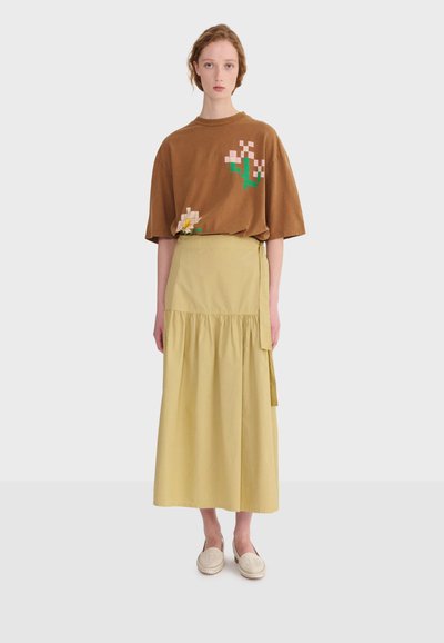 Žena nosi smeđu preveliku majicu s pikseliziranim otiscima cvijeća, bež suknju visokog struka s preklopom i kremaste tenisice bez vezica.