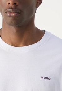 Bílý tričko s kulatým výstřihem vyrobené z bavlny, se malým purpurovým logem "HUGO" na levé části hrudi. Hladký materiál, krátké rukávy.