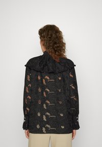 Blouse noire avec détails complexes en dentelle, col à volants et manches longues. Présente des motifs floraux avec sections ajourées et accents de boutons.