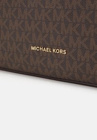 Bolso de tela con estampado marrón que presenta un logo dorado de "Michael Kors". El diseño incluye las iniciales entrelazadas "MK" con un acabado texturizado.