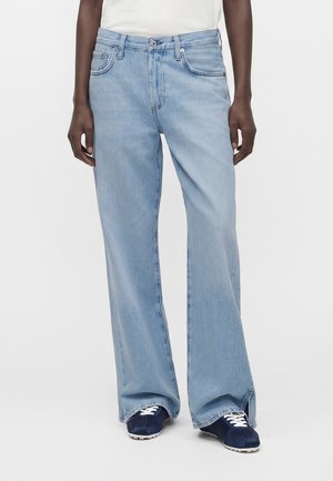 Personne portant un jean bleu clair à jambes larges avec des poches avant et des baskets bleu marine avec des lacets blancs, debout sur un fond blanc.