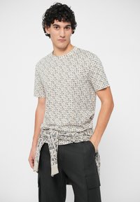 Joven con cabello rizado que lleva una camiseta de color beige con un patrón y un suéter a juego atado alrededor de pantalones cargo negros, de pie contra un fondo blanco.