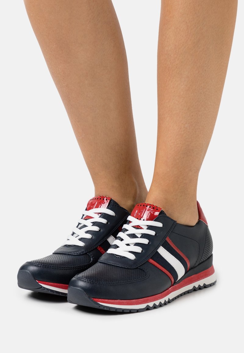Personne portant des baskets sportives noires, rouges et blanches avec des lacets blancs, debout sur un fond blanc.
