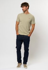 Tricou din tricot texturat, de culoare khaki deschis, cu mâneci scurte și un mic logo roșu; asortat cu blugi slim-fit de culoare albastru închis și adidași bej.