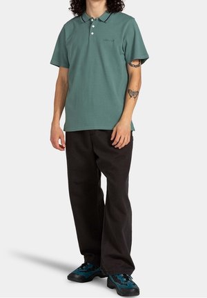 Polo en coton vert avec col et trois boutons, associé à un pantalon large noir et des chaussures avec des accents turquoise.