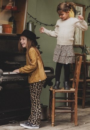 Twee kinderen interacteren in een rustieke kamer. Eén speelt op een zwarte piano, gekleed in een mosterdkleurige trui en bloemenbroek. De ander staat op een kruk in een witte trui en een gemusterde rok.
