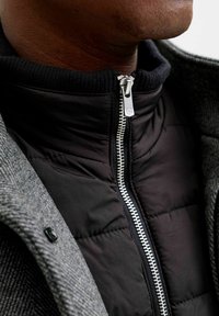 Zwarte pufferjack met een hoge kraag, zilveren rits en een gestructureerde grijze buitenlaag. Voorzien van ribgebreide manchetten en zichtbare stikseldetails.