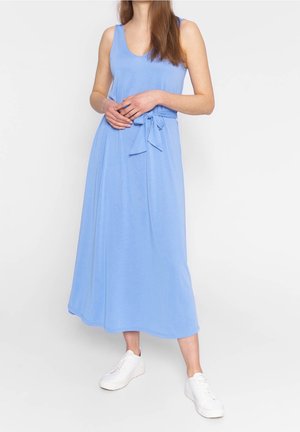 Vrouw in een mouwloze lichtblauwe midi-jurk met een strikceintuur en witte sneakers, staand voor een effen achtergrond.