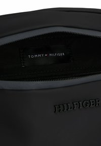 Busta nera con zip caratterizzata da un esterno liscio, fodera in tessuto e un'etichetta con la scritta "TOMMY HILFIGER". Logo in rilievo discreto sulla parte anteriore.