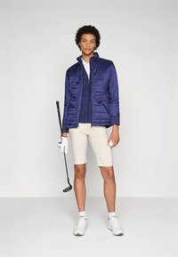 Kobieta stoi, trzymając kij golfowy i piłkę, ubrana w granatową kurtkę, białe szorty, białą rękawiczkę i białe sneakersy na jednolitym tle.