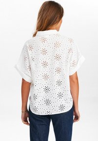 Camicia bianca ricamata con maniche corte, caratterizzata da ritagli floreali e una vestibilità rilassata. La vista posteriore mette in evidenza motivi e trame intricati.
