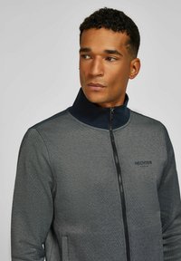 Man met kort krullend haar, gekleed in een grijze ritsjacket met een donkere kraag, die iets naar rechts kijkt tegen een effen achtergrond.