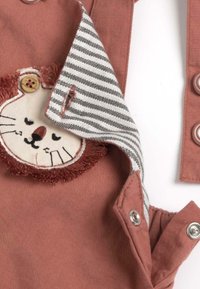 Salopette en tissu couleur rouille avec un appliqué de visage de lion présentant une crinière texturée. La doublure intérieure est rayée en gris et blanc avec des boutons-pression.