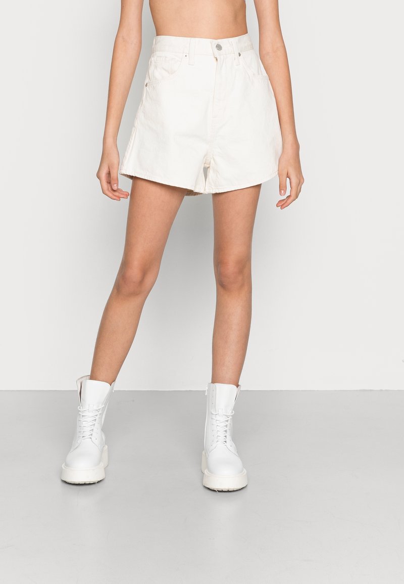 Vita denimshorts med hög midja, framfickor och avslappnad passform, i kombination med vita snörade kängor. Slät textur och rent design.