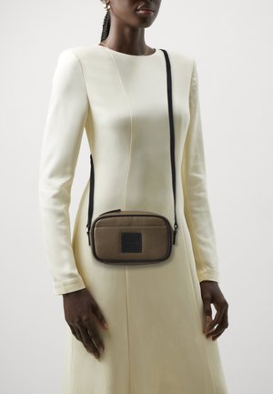 Bolso cruzado marrón con textura, cierre de cremallera, detalles en negro y una correa ajustable, llevado por una figura con un vestido largo blanco.