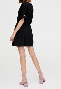 Czarna krótka sukienka z elastyczną talią, podwiniętymi rękawami z detalem na guziki, zestawiona z lawendowymi sandałami na klockowym obcasie. Gładka faktura tkaniny.