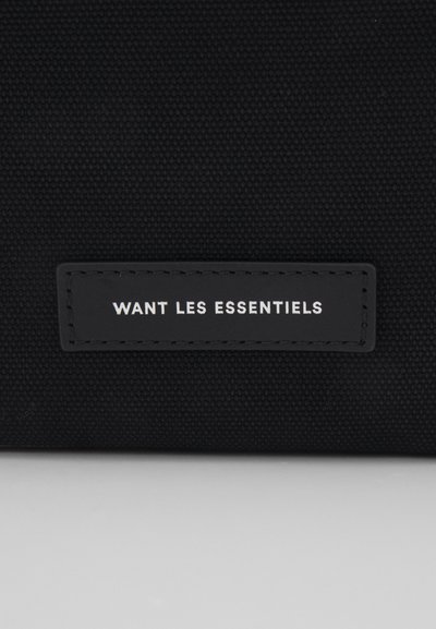 Borsa di stoffa nera con un'etichetta rettangolare con il testo "WANT LES ESSENTIELS" in bianco. Superficie testurizzata con dettagli di cucitura.