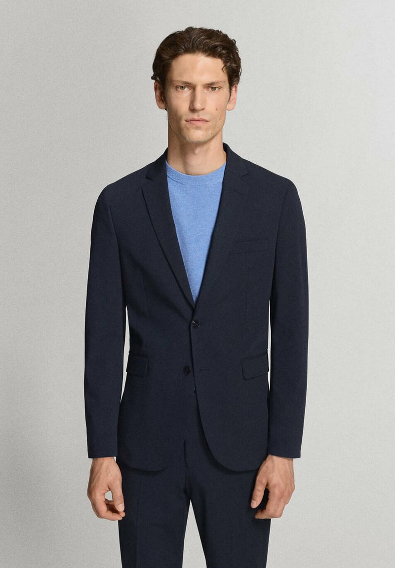 Homme aux cheveux courts et bouclés portant un costume bleu marine foncé et un pull bleu clair à col rond, debout devant un fond uni gris clair.