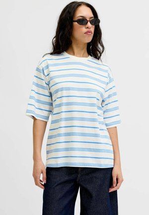 Kobieta nosząca czarne owalne okulary przeciwsłoneczne, białą oversize koszulkę z jasnoniebieskimi poziomymi paskami i ciemnoniebieskie dżinsy, stojąca na białym tle.