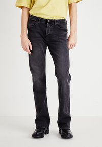 Črne denim hlače s ravnim krojem, ki imajo vidne žepke in rahlo zbledelost. Zesafte s lahkim rumenim srajcem in črnimi čevlji.