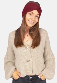 Cardigan en tricot beige avec trois boutons dorés, présentant des manches larges et une coupe courte, associé à un bandeau texturé bordeaux.