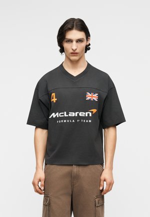 Giovane uomo con capelli scuri indossa una maglietta nera del McLaren Formula 1 Team con il numero 4 e una toppa con la bandiera del Regno Unito, abbinata a pantaloni cargo marroni.