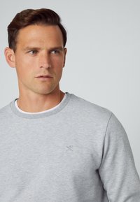 Grauer Pullover mit Rundhalsausschnitt und feiner Textur. Mit einem kleinen gestickten Emblem auf der Brust, wobei ein weißes Unterhemd sichtbar ist.