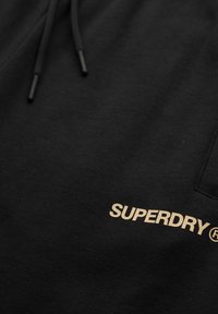 Zwarte hoodie met een beige "SUPERDRY®" logo gedrukt nabij de zak en zwarte trekkoorden met afgewerkte uiteinden.