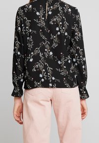 Blusa nera con un motivo floreale dai colori tenui; presenta maniche lunghe, una scollatura a chiave sul retro e polsini leggermente arricciati sopra pantaloni rosa.