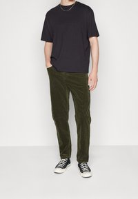 Zwarte T-shirt met korte mouwen, gecombineerd met groene corduroy broeken. De broeken hebben een gestructureerd oppervlak en zijn iets taps toelopend. Zwarte sneakers maken de look compleet.