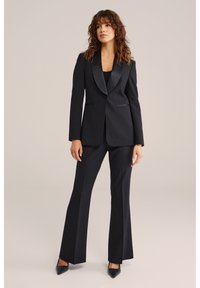 Svart skräddarsydd kostym med satinfodrad kavajslag, smal passform på kavajen med fickor och utsvängda byxor. Kombinerad med svarta spetsiga klackar.
