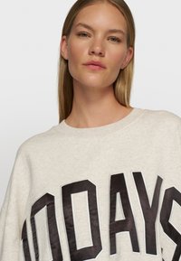 Hellgraues, übergroßes Sweatshirt mit schwarzem "100 DAYS"-Text, ausgestattet mit einem Rundhalsausschnitt und weichem, strukturiertem Stoff.