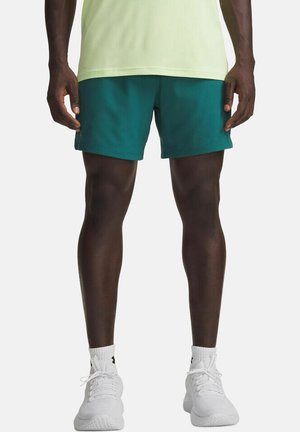Man draagt turquoise sportshorts, witte enkelsokken en witte hardloopschoenen, staand met armen ontspannen langs het lichaam tegen een effen achtergrond.
