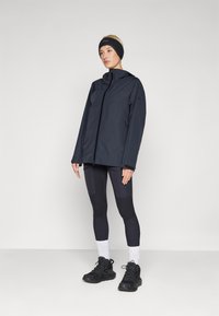 Jachetă neagră rezistentă la apă, cu închidere cu fermoar și glugă, purtată cu leggings negri și pantofi sport negri, completată cu o bandă pentru cap.